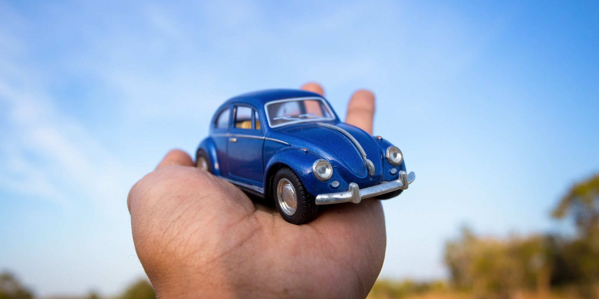 Eine Hand hält ein blaues Spielzeugauto vor blauem Himmel im Freien.